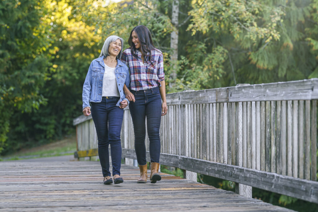 A vibrant senior woman of Asian descent links arms with her mixed race adult daughter as they walk across a footbridge in a natural parkland area. 