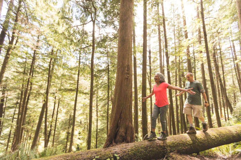 Cute senior couple explore the Pacific Northwest together on a day hiking trip. They are walking across a log in the forest. She is in front and they have their arms out for balance as they inch across.